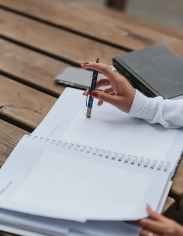 Mão a apontar para calendário mensal num planeador, com caneta e caderno sobre mesa de madeira, repr