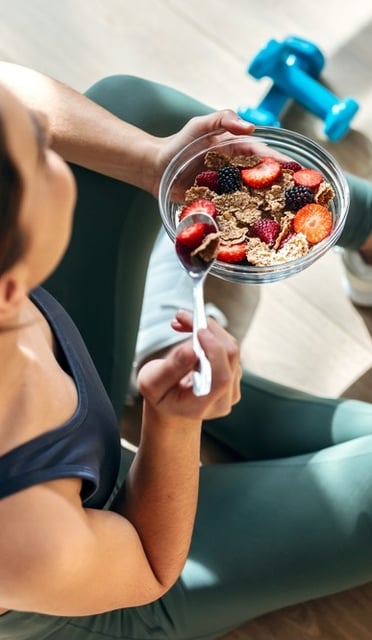 A woman eating cereal and fruits from a bowl, with small weights and a bottle of water on the floor of the gym studio