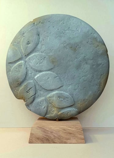 A round plate-like sculpture depicting leaf-shaped motifs, painted in blue greys, on oak plinth.