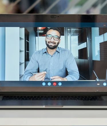 A laptop screen showing a video call with a man in a business shirt holding a pen