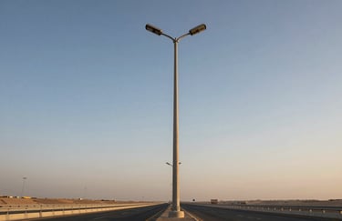A photograph of a tall, industrial-grade street light pole standing prominently along a modern highway in the Middle Eastern / Gulf region, during the golden hour.