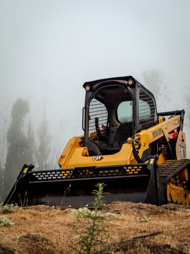 A yellow Caterpillar compact track loader parked on a foggy construction site with a front bucket attachment.