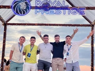 Five young men smiling under a Royal Prestige corporate event sign with outdoor lighting at dusk.
