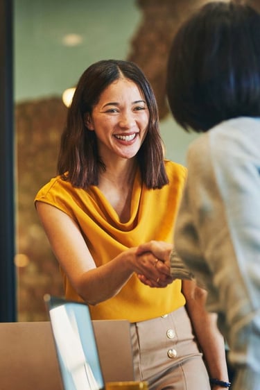 Smiling business professional in a yellow blouse shaking hands with a client in a modern office.