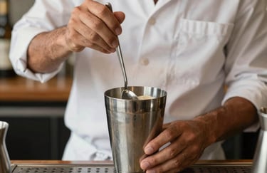 Artisan beverage preparation. A pair of hands in professional attire stirring a drink with a long silver spoon in a mixing glass. South American setting. Natural light.
