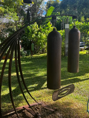 Rustic metal outdoor wind chimes hanging in a sunny tropical garden with lush green foliage.