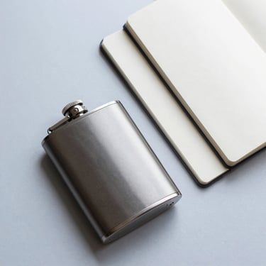 An artistic flat-lay of lifestyle essentials, including a sleek matte flask and a modern notebook, on a pale blue-grey surface, bright and airy lighting.