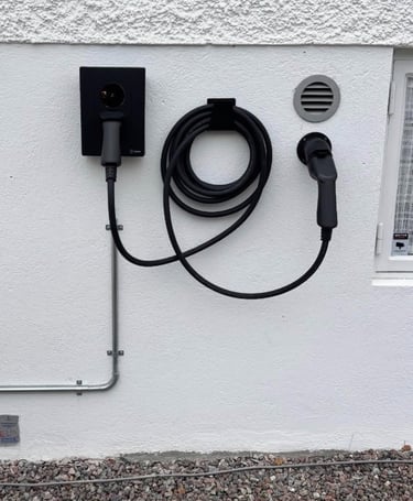 A wall-mounted black EV home charging station with a coiled cable and connector on a white exterior wall.