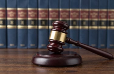 A wooden legal gavel resting on its sound block on a polished desk. The background is a wall of classic law books in soft focus. Colors: dark slate blue and wood tones.