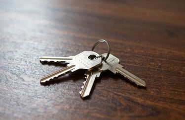 A professional photography shot of a set of silver house keys resting on a polished dark wood table with soft sunlight streaming in, North American / US style.