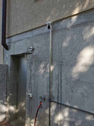 A white outdoor security camera mounted on a concrete wall with metal conduit and a wall lantern.