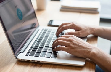 A close-up of hands typing on a modern laptop in a bright, sunlit cafe in North America. A smartphone next to the laptop displays the Qorabot notification icon. The scene is approachable and intelligent with Sky White tones.