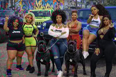 Six women in Jeep apparel posing with two black Cane Corso dogs in front of colorful urban graffiti and SUVs.