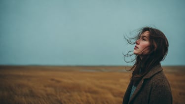 A pensive woman in a wool coat stands in a windswept field under a moody, overcast autumn sky.