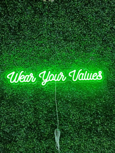 grünes LED Neon Schild mit Schriftzug „Wear Your Values“ auf grüner Pflanzenwand