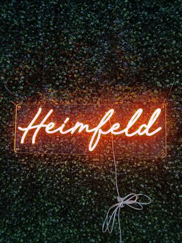 oranges LED Neon Schild mit Schriftzug „Heimfeld“ auf grüner Pflanzenwand