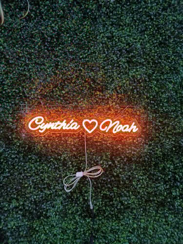 oranges LED Neon Schild mit Schriftzug „Cynthia ♥ Noah“ auf grüner Pflanzenwand