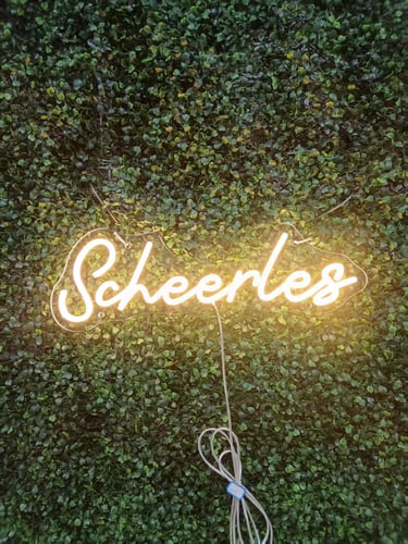 warmweißes LED Neon Schild mit Schriftzug „Scheerleo“ auf grüner Pflanzenwand