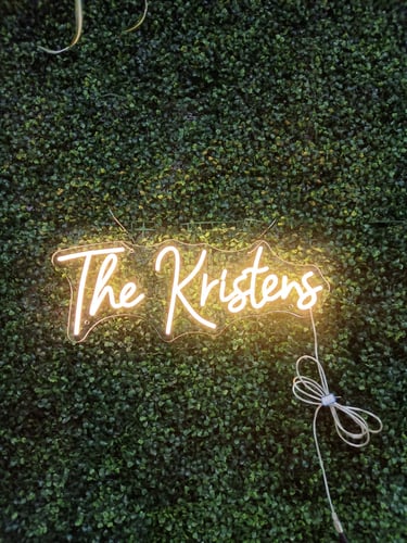 warmweißes LED Neon Schild mit Schriftzug „The Kristens“ auf grüner Pflanzenwand