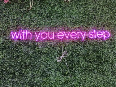 pinkes LED Neon Schild mit Schriftzug „with you every step“ auf grüner Pflanzenwand