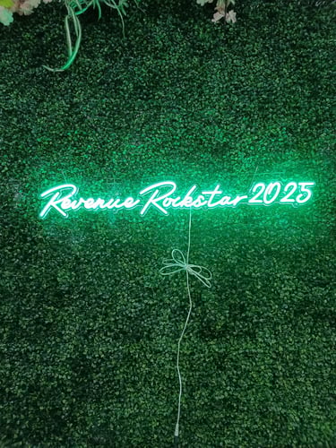 grünes LED Neon Schild mit Schriftzug „Revenue Rockstar 2025“ auf grüner Pflanzenwand bei Event