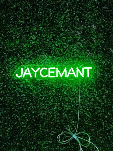 grünes LED Neon Schild mit Schriftzug „JAYCEMANT“ auf grüner Pflanzenwand Hintergrund