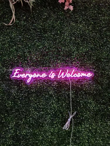 pinkes LED Neon Schild mit Schriftzug „Everyone is Welcome“ auf grüner Pflanzenwand Hintergrund