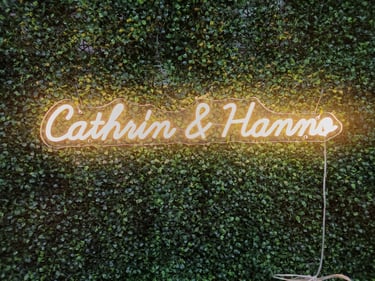 Warm Weißes Neon Schild „Cathrin & Hanno“ personalisierter LED Neon Schriftzug für Hochzeit auf grüner Pflanzenwand