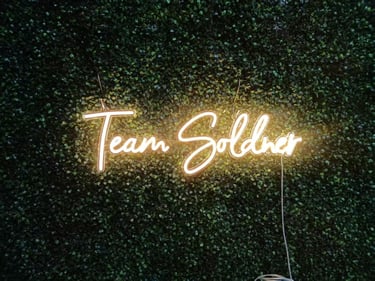 Warm weisses Neon Schild „Team Soldner“ personalisierter LED Neon Schriftzug auf grüner Pflanzenwand