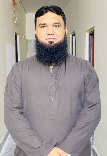 A bearded man wearing a traditional brown kurta stands in a hallway.