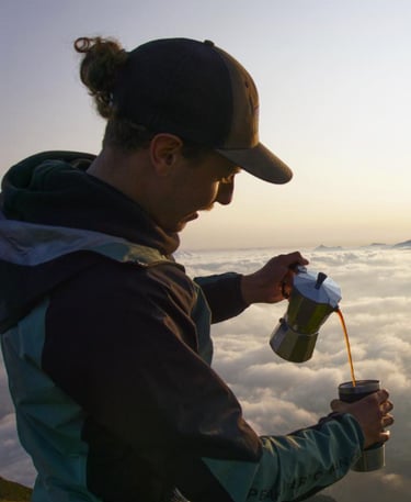 A man pours coffee from a moka pot into a mug while standing above the clouds during a mountain sunrise.