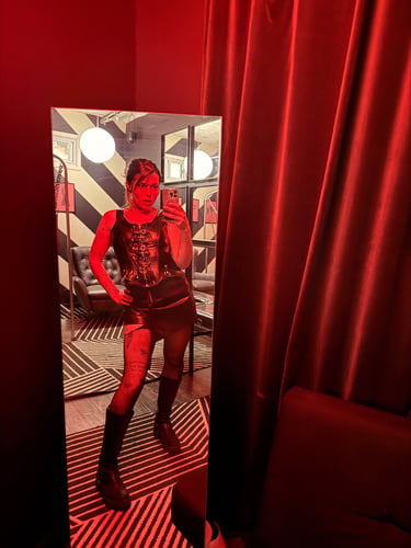 Dominatrix wearing leather vest, mini skirt and knee high boots, posing in a red lit mirror 