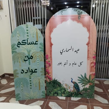 استاندات استقبال العيد بتصاميم متنوعة، أحدها بنقش النخيل وعبارة عساكم من عواده، والآخر بتصميم قنطرة 