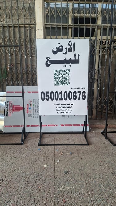 لوحة إعلانية عقارية بيضاء مكتوب عليها الأرض للبيع مع كود QR ورقم تواصل مثبتة على حامل معدني أسود متي
