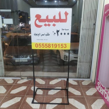 لوحة عقارية بيضاء مكتوب عليها للبيع باللون الأحمر مع حامل معدني أسود أمام معرض سيارات ومكتب عقار