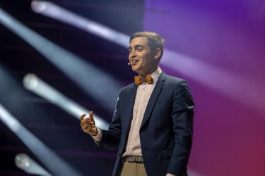 A smiling male keynote speaker in a blue suit and bow tie presenting on a stage with stage lights.