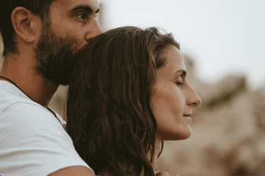 A bearded man kisses a woman with wavy hair on the forehead during a romantic outdoor sunset session.