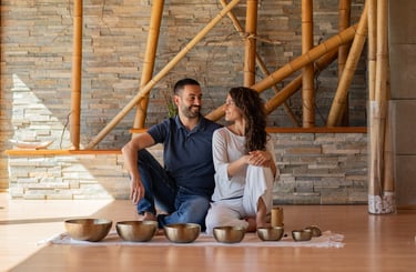 A smiling couple sits on a wooden floor behind brass Tibetan singing bowls for a sound healing session.