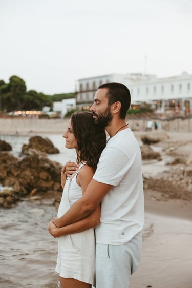 A romantic couple in white clothes hugging on a rocky beach at sunset.
