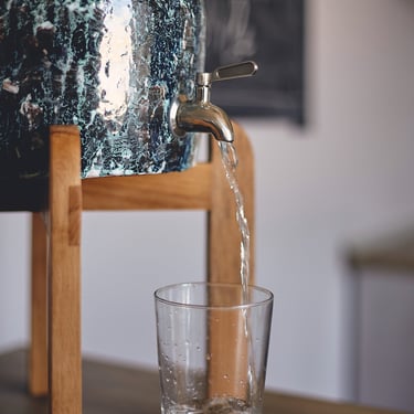 Сeramic dispenser with sea-wave textured glaze, stainless steel spigot, and sturdy oak legs, pouring water into a glass
