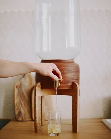 Rustic ceramic dispenser on oak tabletop stand, water pouring from spigot into glass with lemon slice, hand adjusting tap