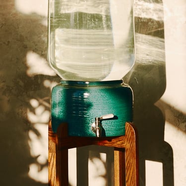 Elegant ceramic water dispenser with glass bottle, placed by loft-style wall with sunlight reflections and plant shadows