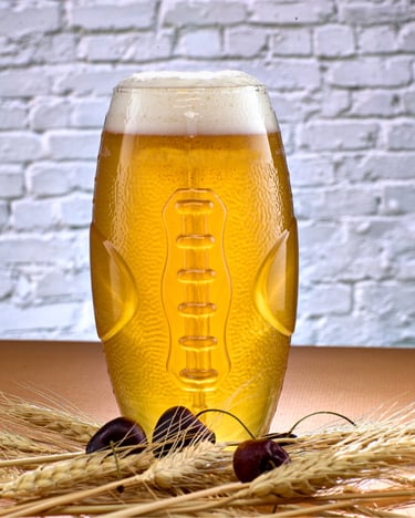 Samuel Adams Cherry Wheat beer in a Football shaped glass, with 3 cherries and wheat in front showcasing light coming through