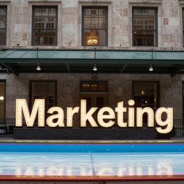 A brightly lit, colorful Toronto sign is displayed in front of a modern building at night. The letters are illuminated in different colors, casting reflections on the ground, with a maple leaf symbol at the end. Silhouettes of people are visible near the sign.