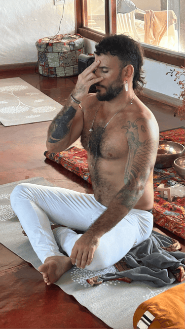 Tattooed man practicing alternate nostril breathing yoga on a mat in a sunlight studio.
