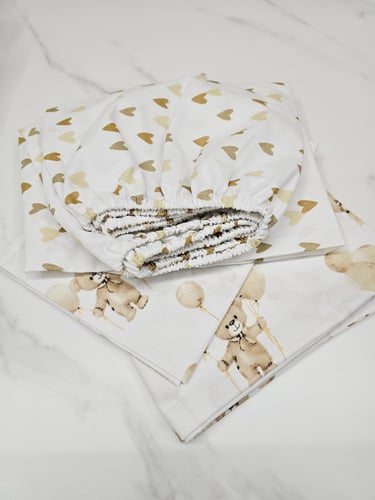 Folded white baby crib bedding set featuring gold hearts and teddy bear balloon patterns.