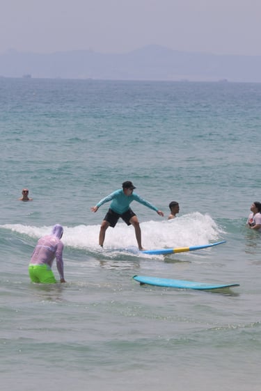 A surfer in a blue rash guard rides a small ocean wave on a surfboard near other swimmers.