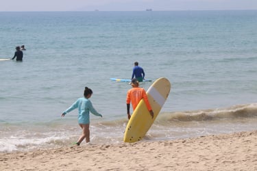A surf instructor in an orange shirt helps a student carry a yellow surfboard into the tropical blue ocean.