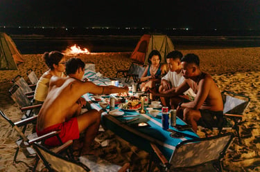 A group of friends enjoying a beach dinner by a bonfire and camping tents at night.