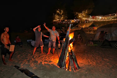 Friends gather around a bright bonfire on a sandy beach at night during a camping trip.